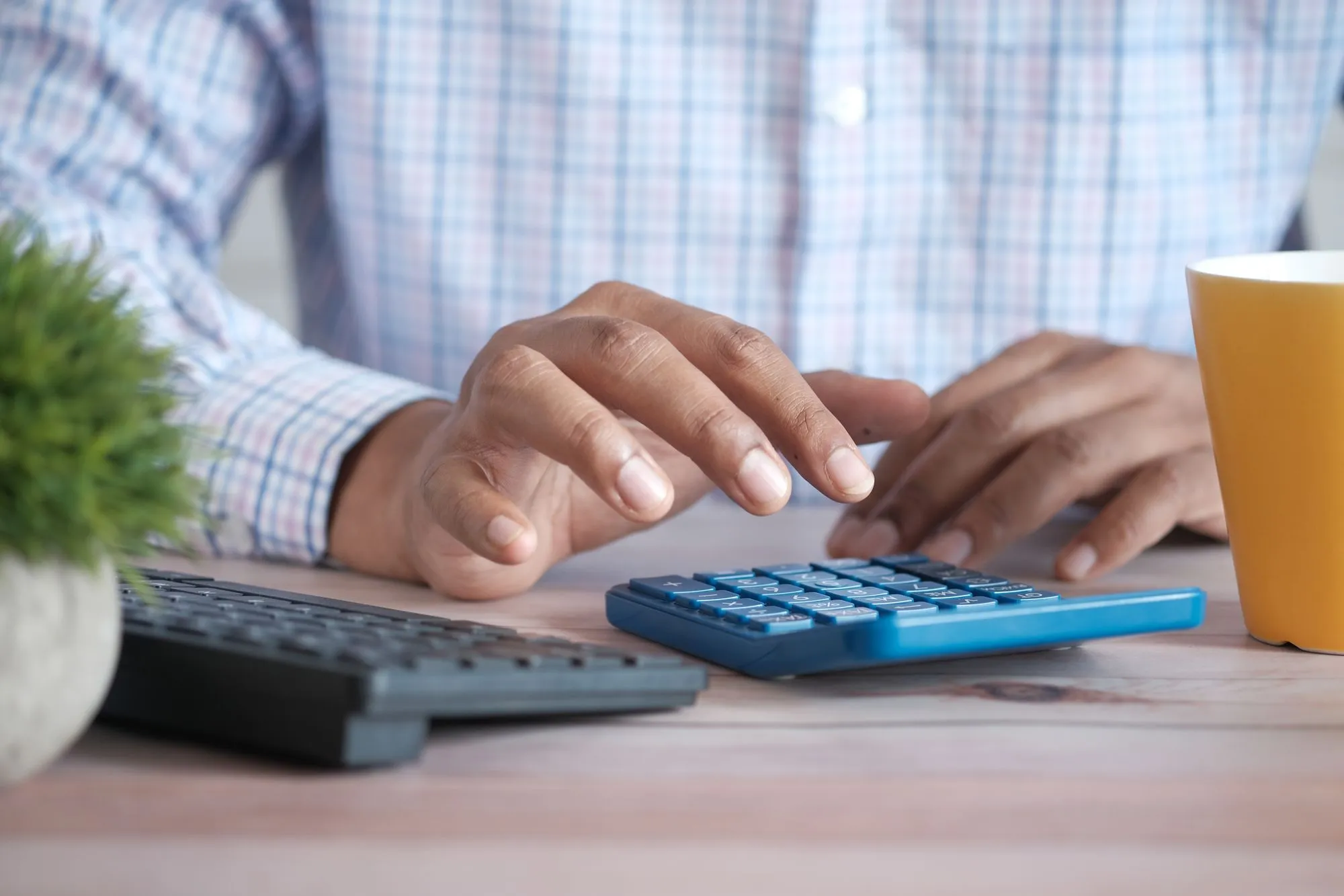 Mãos digitando numa calculadora azul sobre uma mesa de madeira, ao lado de um teclado